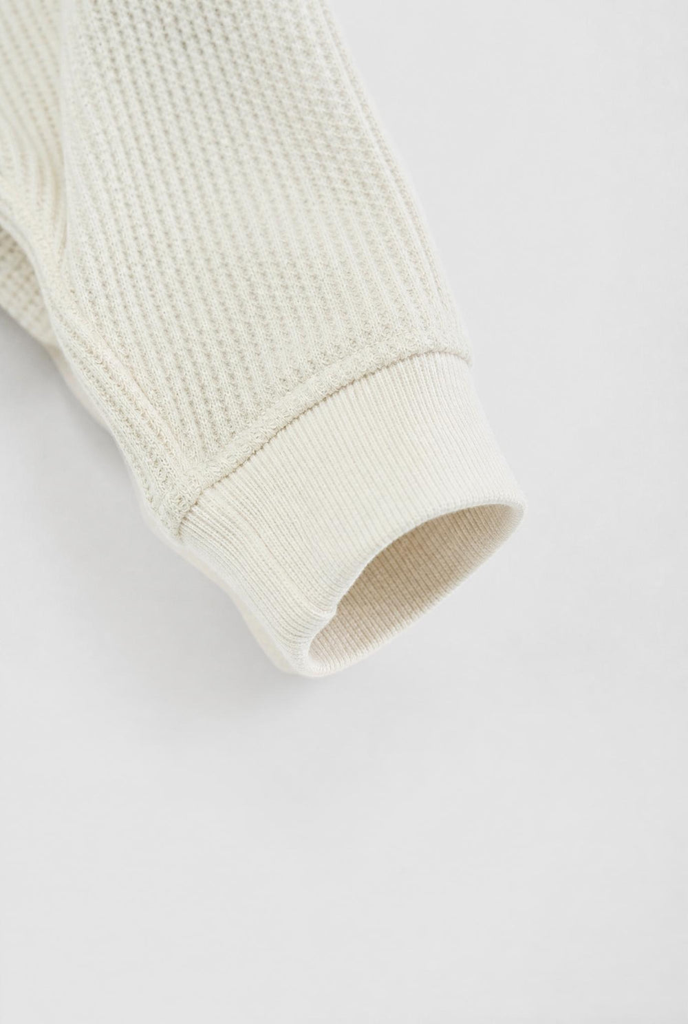 Close-up of a ribbed knit sleeve cuff in a warm off-white or cream color, showing the texture and construction against a plain light background.